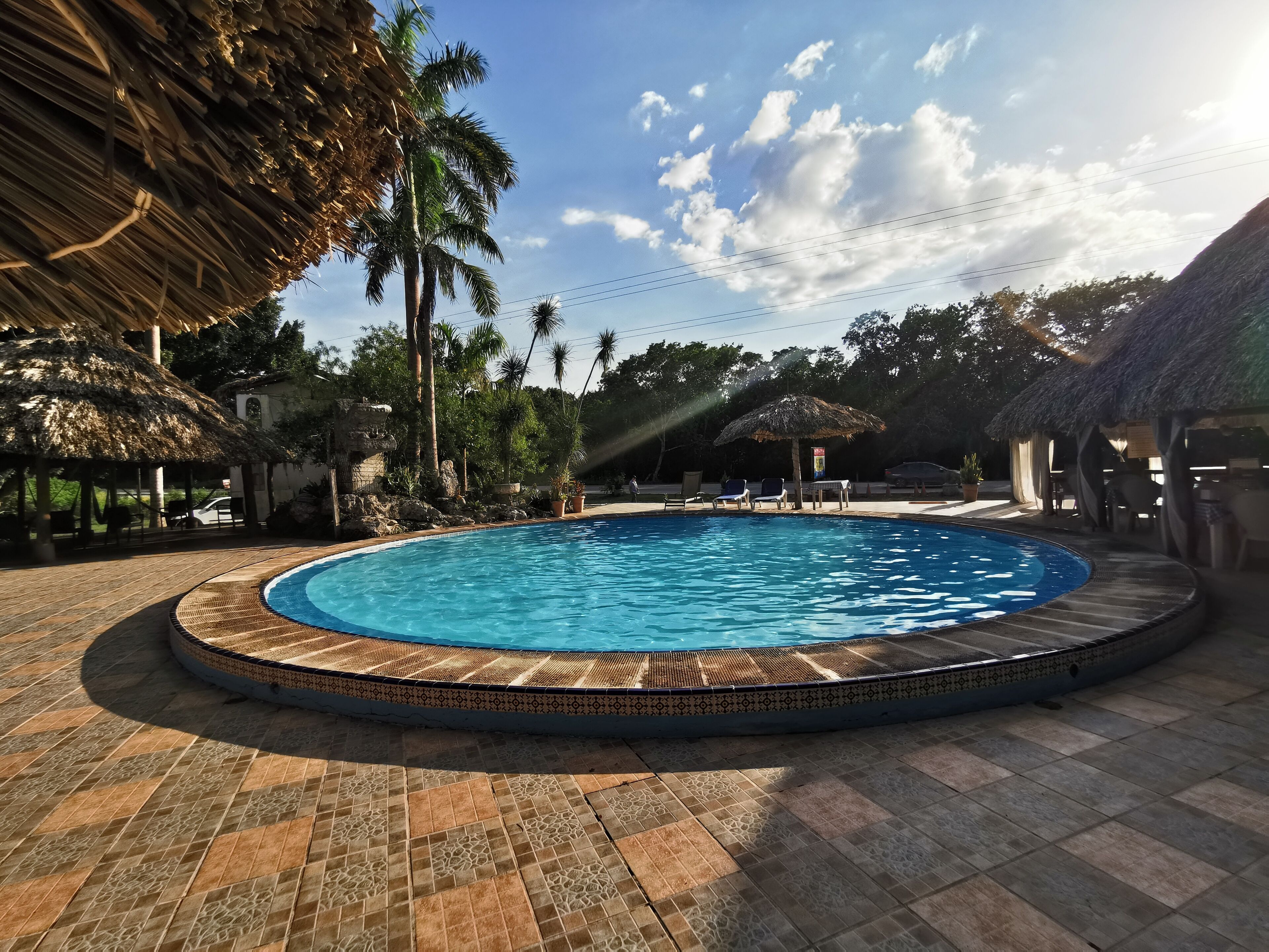 A circular pool with a tiled edge, surrounded by lounge chairs and thatched umbrellas, set against a backdrop of palm trees and a clear sky.