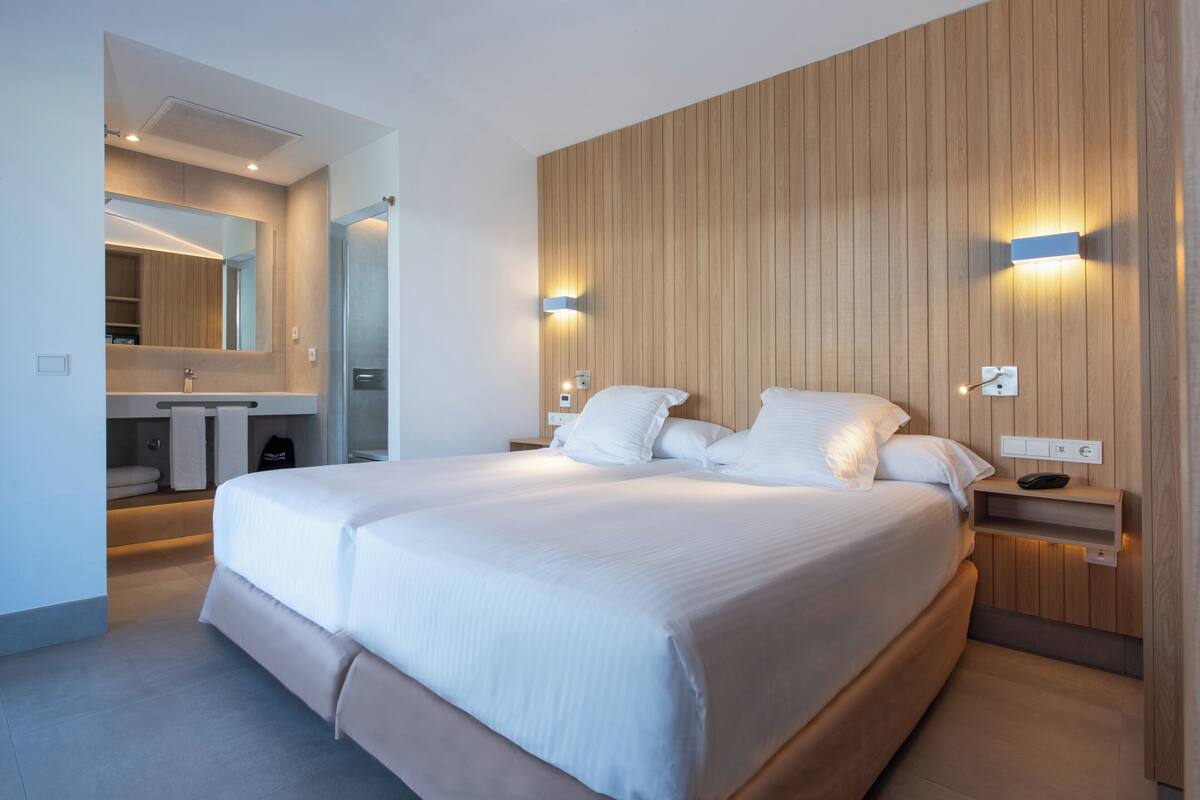 A modern hotel room with a large bed, a wooden panel wall, a built-in desk, and a bathroom with a mirror and towel rack.