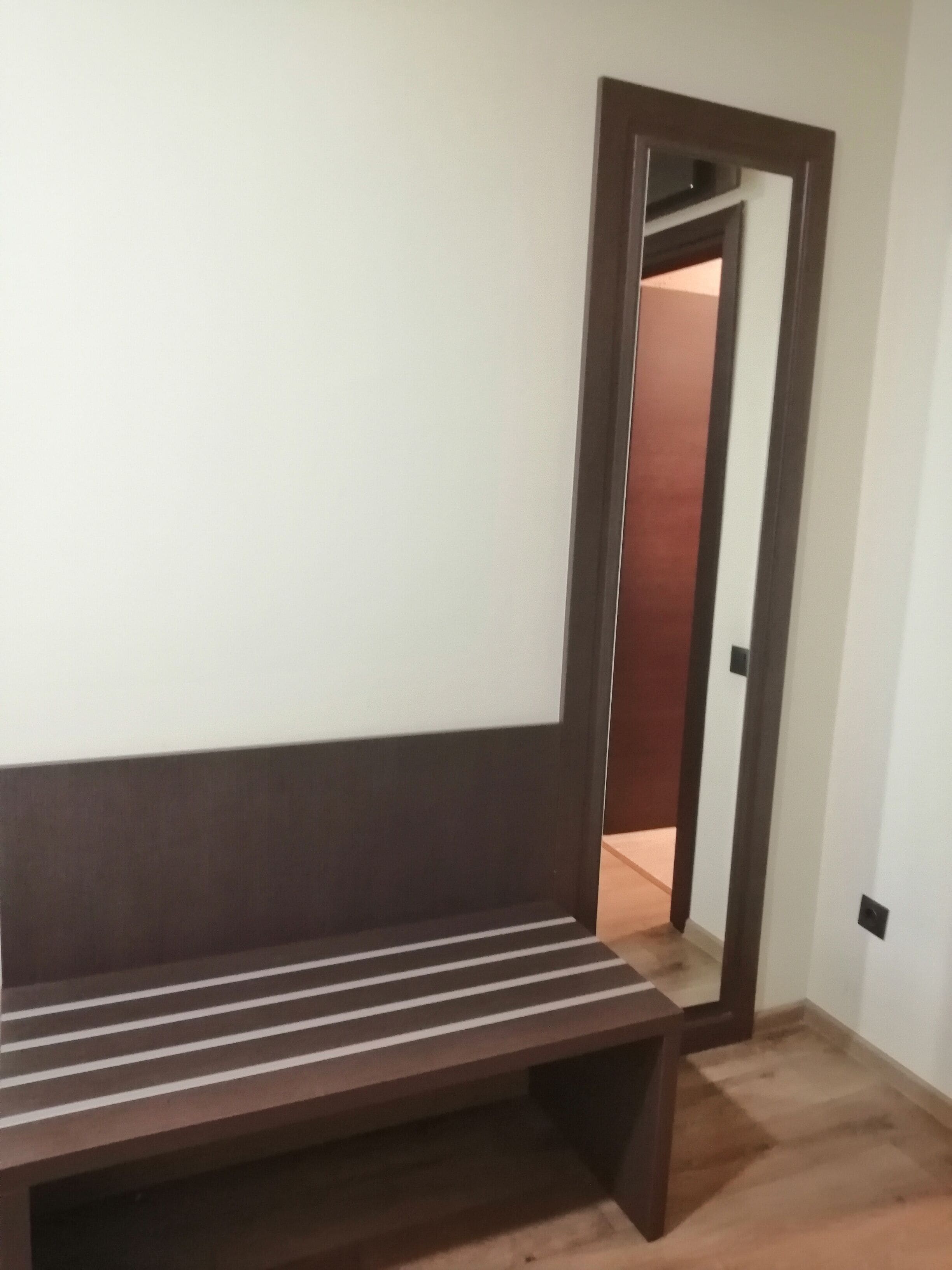 Une pièce avec un banc en bois adossé à un mur blanc, une porte donnant sur une autre pièce et un plancher en bois.