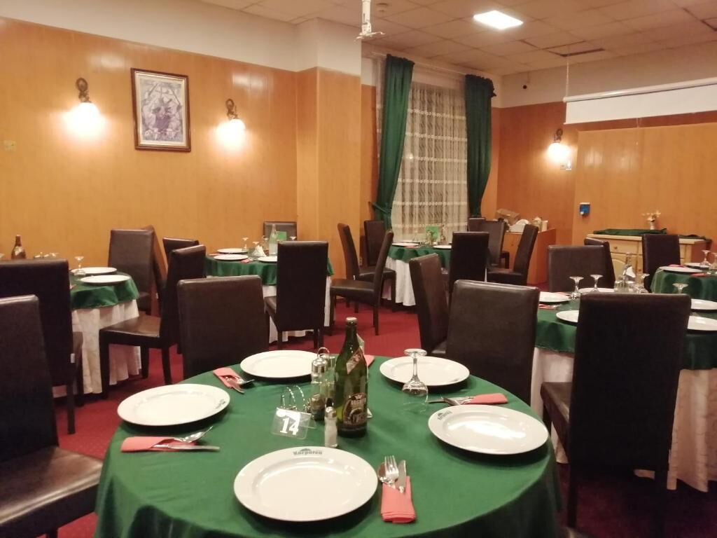 A dining area with round tables set for four, each with white plates, cutlery, and a bottle of wine. The tables are covered with green tablecloths, and the room has wooden paneling and a large window with green curtains.