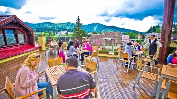 An outdoor terrace with wooden tables and chairs, surrounded by mountains and a cloudy sky. - Hotel Piemonte (Predeal)