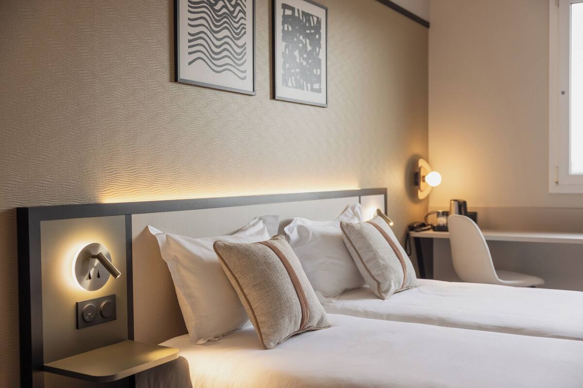 Une chambre d’hôtel avec deux lits, une table de chevet avec une lampe, une chaise et des œuvres d’art accrochées au mur.
