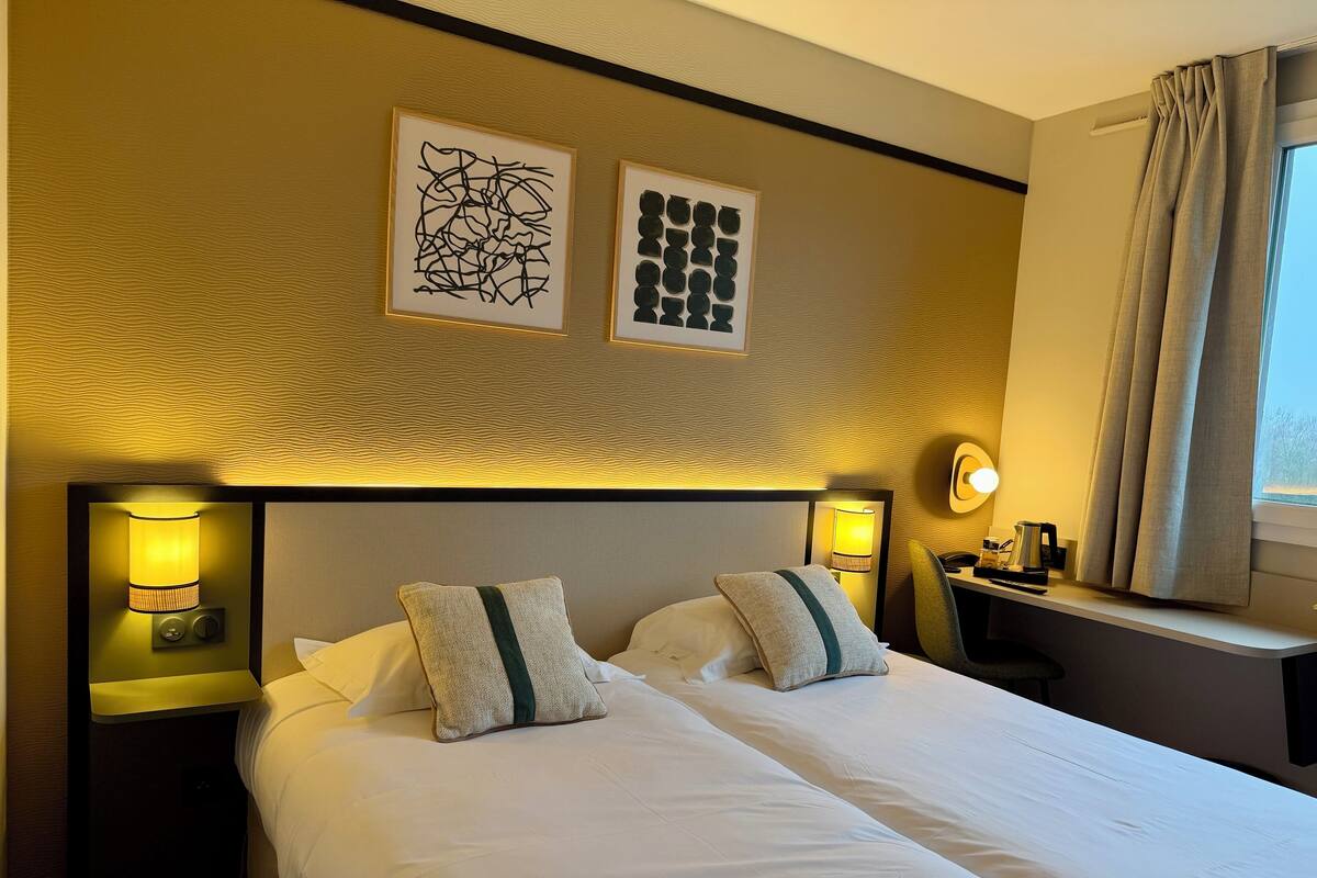 Une chambre d’hôtel avec deux lits, des tables de chevet avec des lampes, un bureau avec une lampe et des œuvres d’art accrochées au mur.