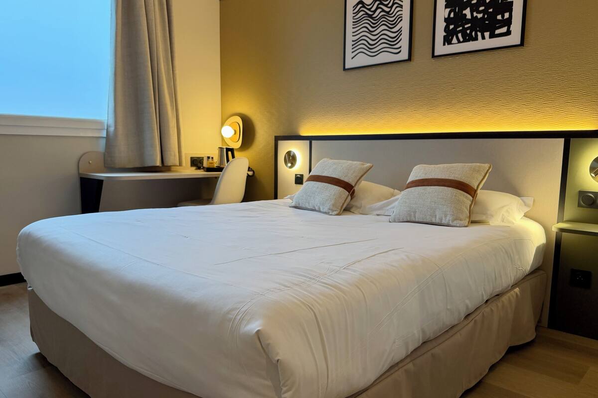 Un grand lit avec du linge de lit blanc, une table de chevet avec une lampe et deux œuvres d’art murales.