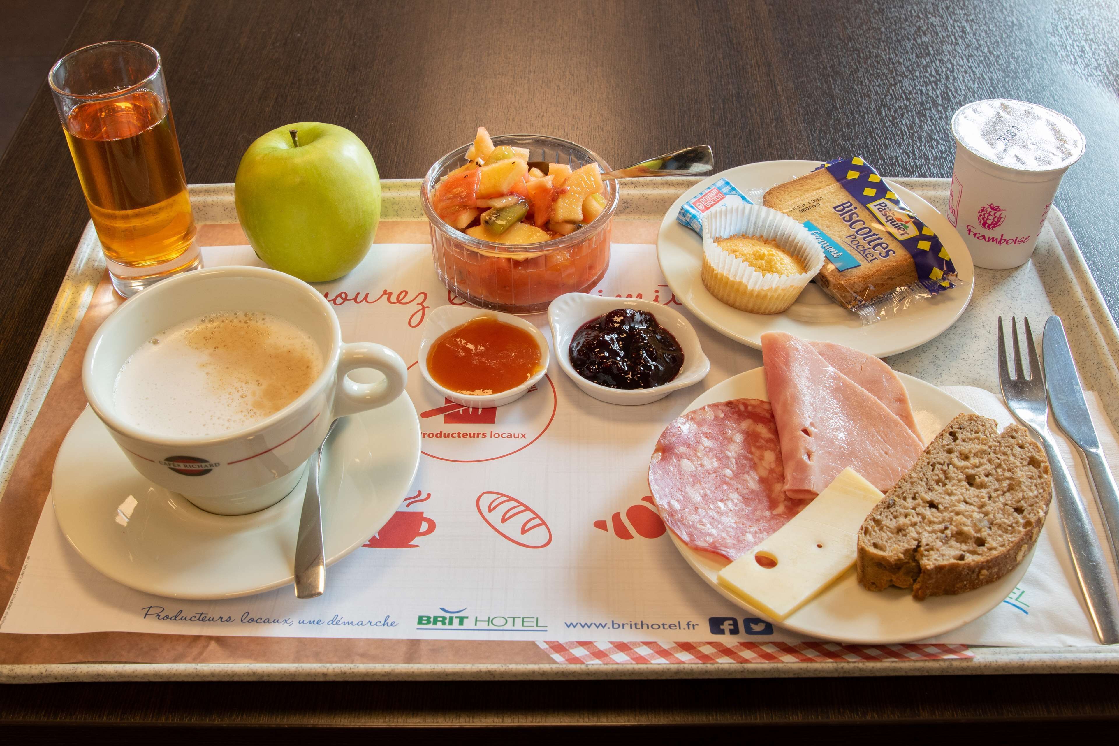 Een dienblad met een maaltijd, inclusief een kopje thee, een appel, een schaal fruit, een bord met ham, kaas, koekjes, een muffin en twee kleine schaaltjes jam.