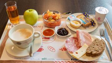 Een dienblad met een maaltijd, inclusief een kopje thee, een appel, een schaal fruit, een bord met ham, kaas, koekjes, een muffin en twee kleine schaaltjes jam.