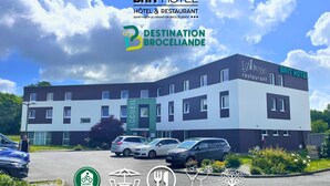 Un bâtiment moderne arborant l’enseigne du Brit Hotel et de son restaurant, avec un espace de stationnement où sont garées plusieurs voitures, ainsi que divers pictogrammes indiquant les services disponibles, tels que le Wi-Fi, le parking et un espace de service ouvert 24 heures sur 24.