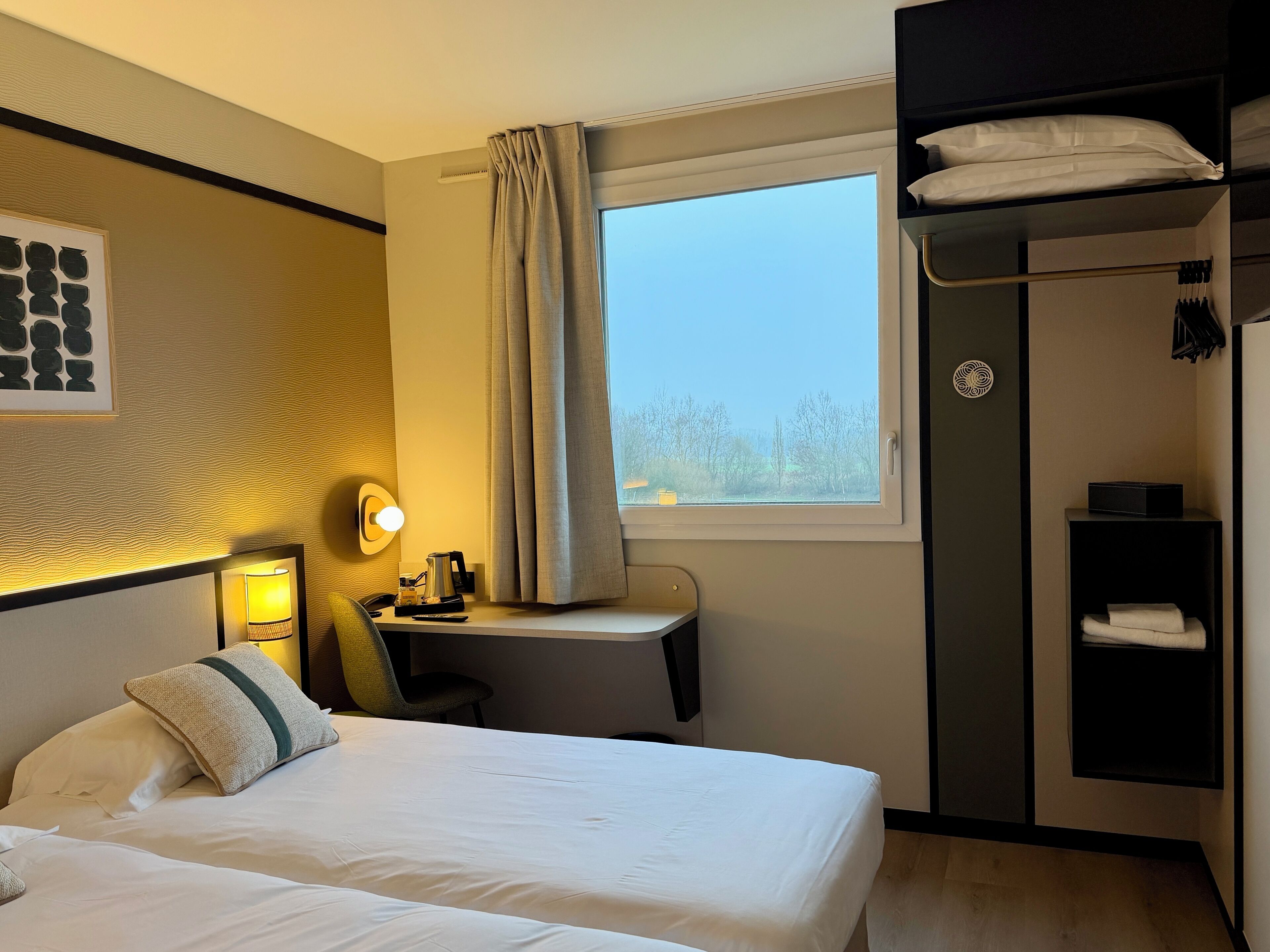Habitación de hotel con cama, mesita de noche, lámpara, escritorio, silla, armario y ventana con cortinas.