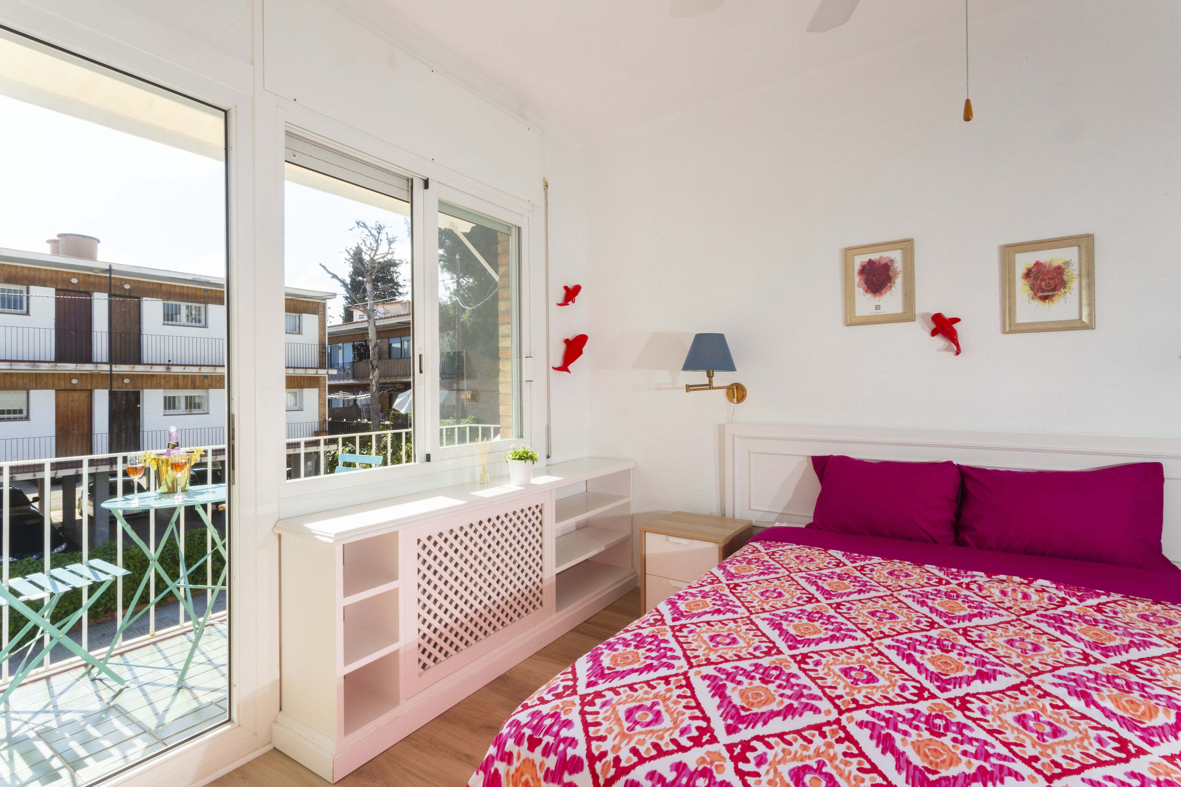 A bedroom with a large window, a balcony, a bed with a patterned bedspread, and a small table with a lamp.