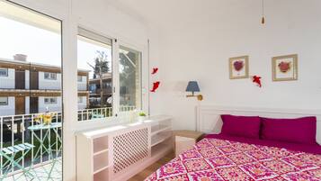 A bedroom with a large window, a balcony, a bed with a patterned bedspread, and a small table with a lamp.