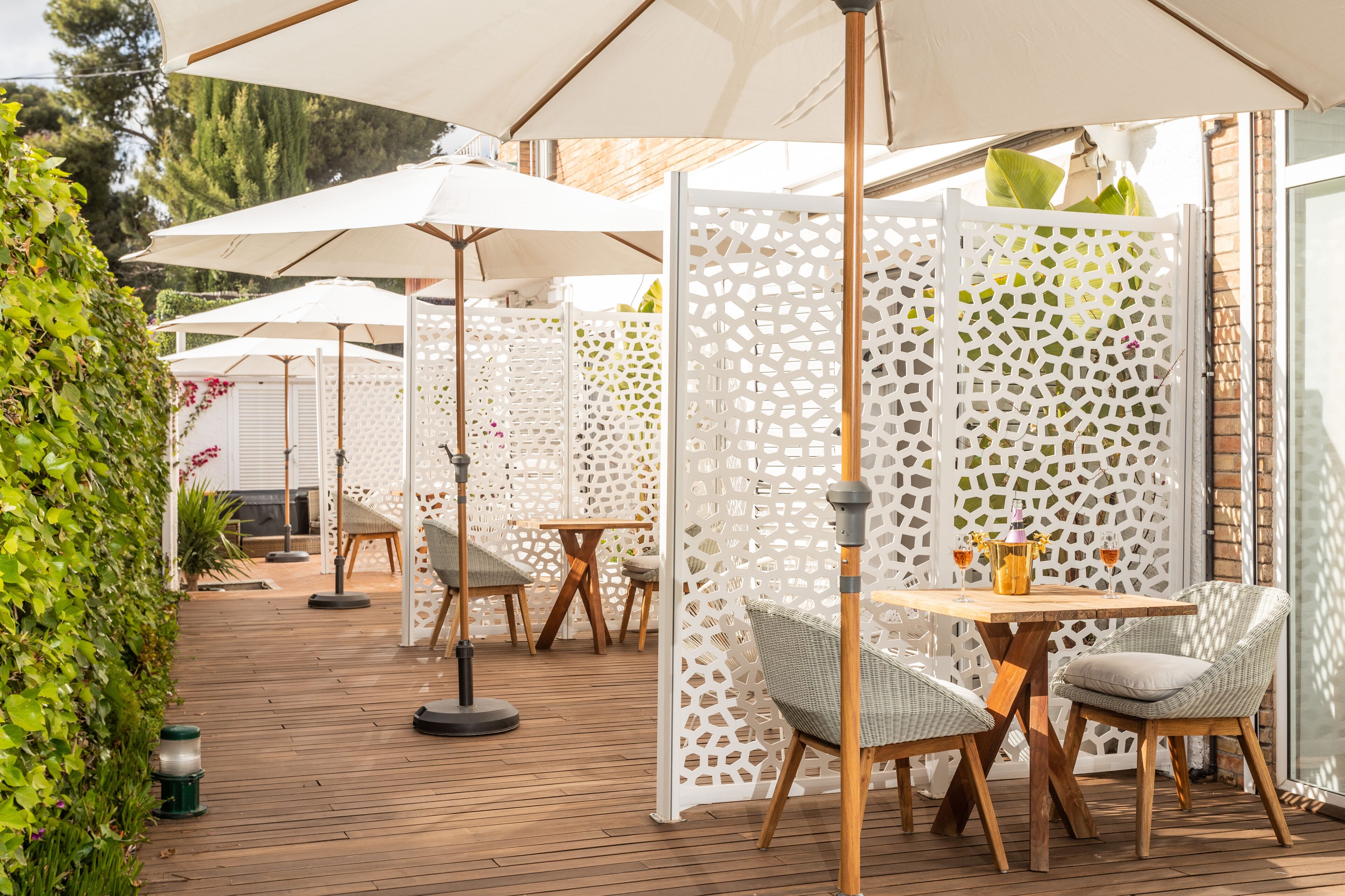Un patio al aire libre con suelo de madera, sombrillas blancas, sillas de mimbre y una mesa con un florero y bebidas.