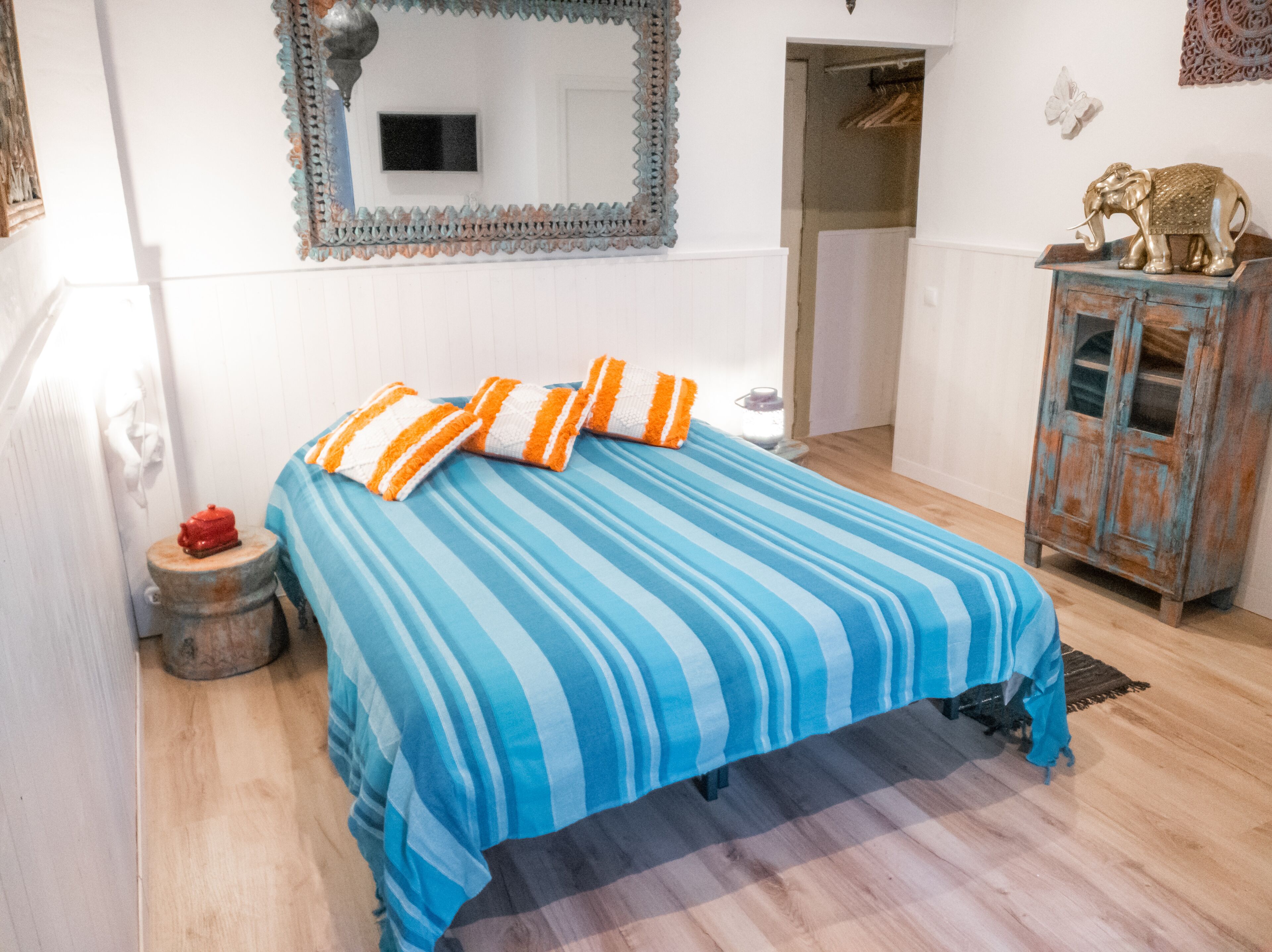 Uma cama com roupa de cama listrada, uma pequena mesa de cabeceira com um objeto vermelho, um espelho na parede e um armário com um elefante decorativo.