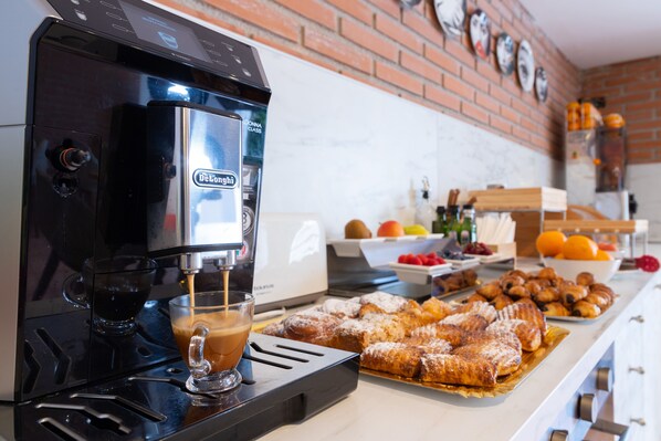 A coffee machine dispensing espresso into a cup, a tray of pastries, and a fruit bowl on a kitchen counter.