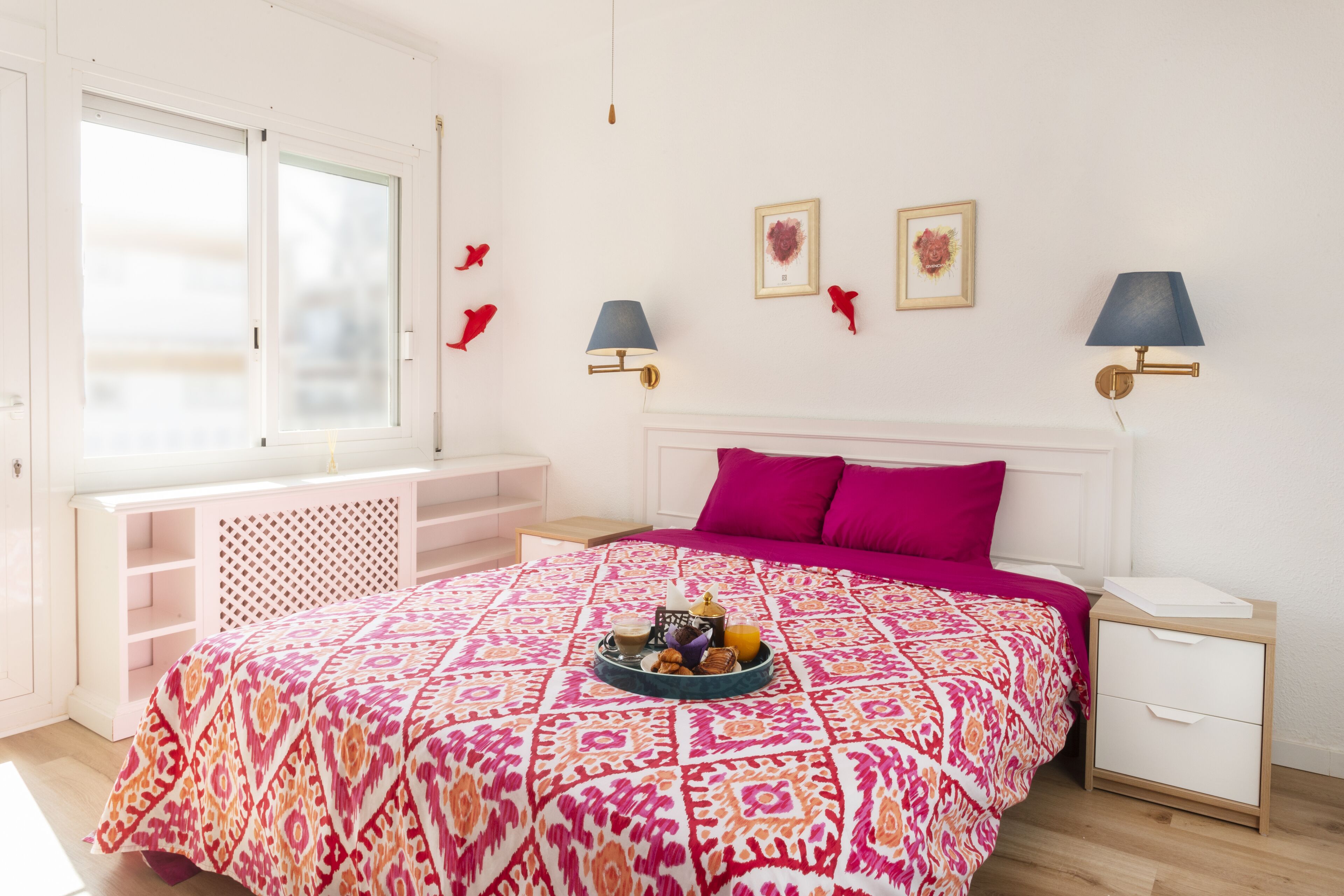 A bedroom with a patterned bedspread, a bedside table with a lamp, a window with blinds, and a small shelf.