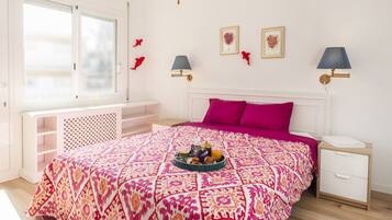 A bedroom with a patterned bedspread, a bedside table with a lamp, a window with blinds, and a small shelf.