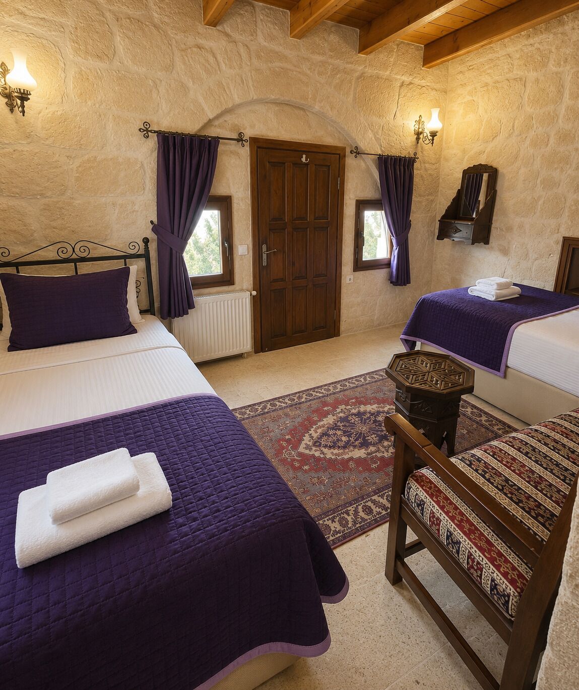 Una habitación con dos camas, una silla de madera, una mesita y un muro de piedra.