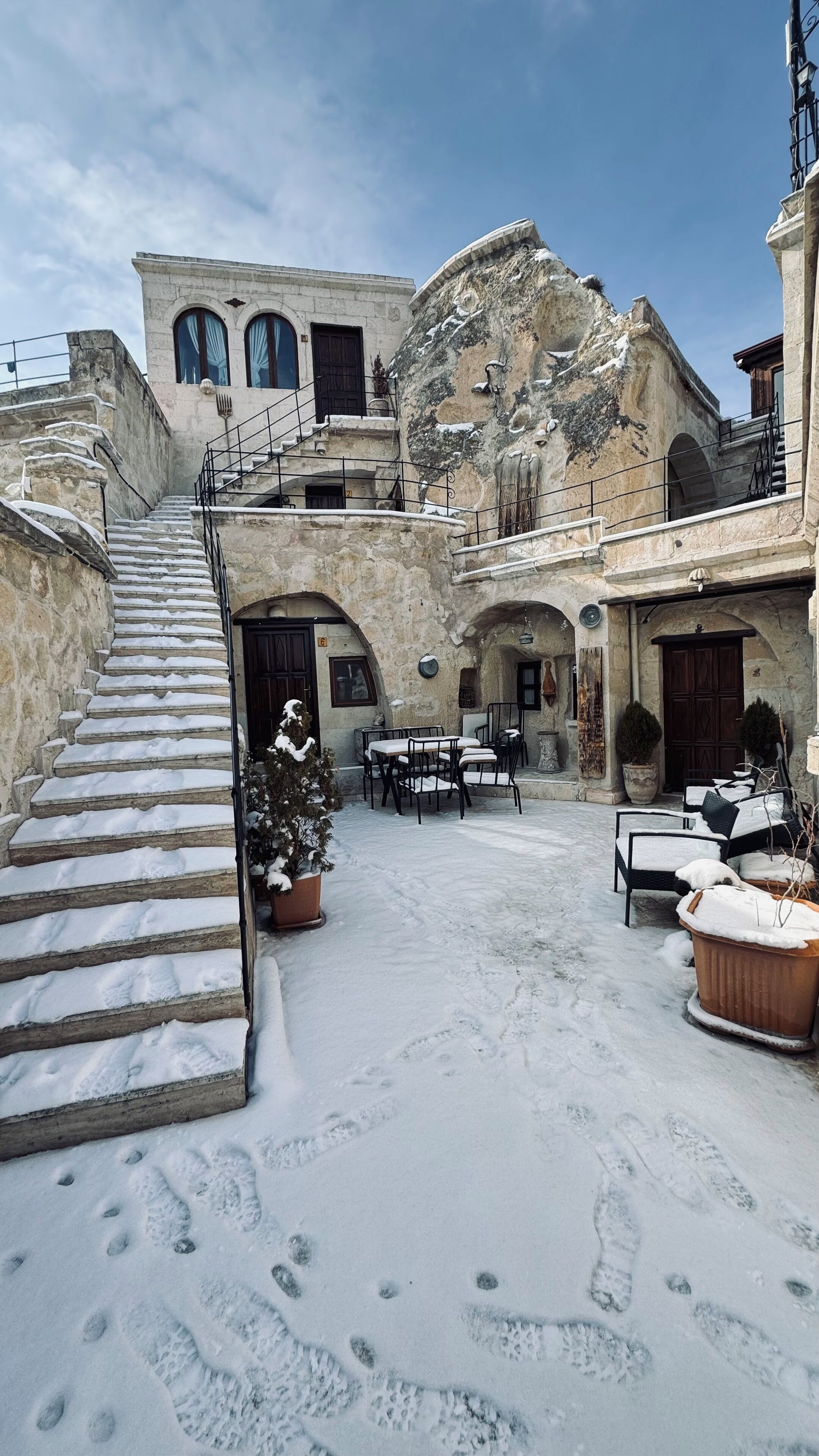 A courtyard with a stone staircase leading to a building with multiple windows and doors. There are outdoor tables and chairs, and a potted plant.
