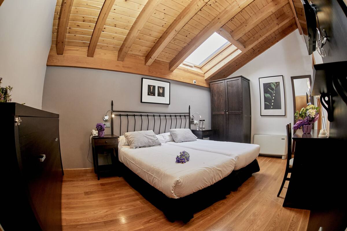 A bedroom with a large bed, wooden ceiling beams, a skylight, a wardrobe, and a desk with a chair.