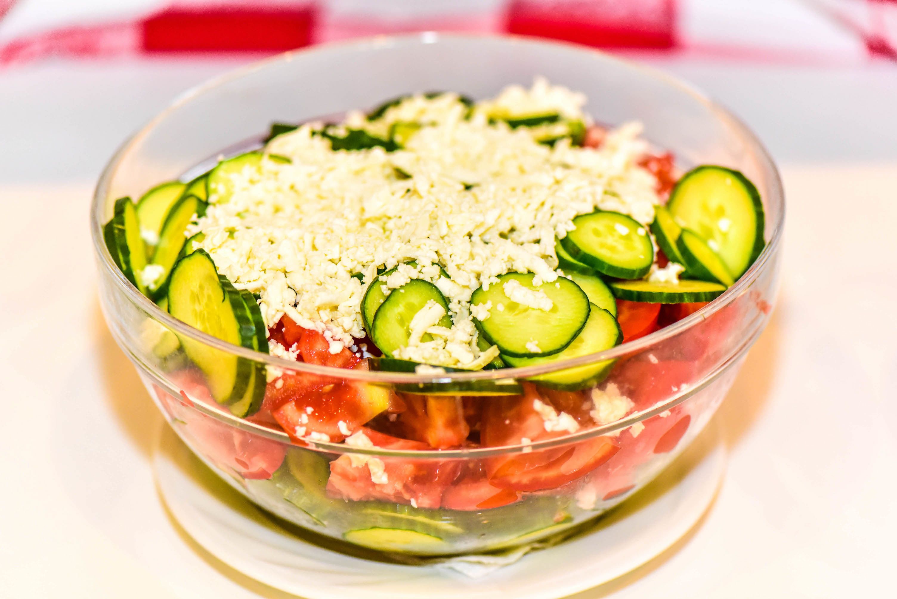 A clear glass bowl containing a salad with cucumbers, tomatoes, and shredded cheese.