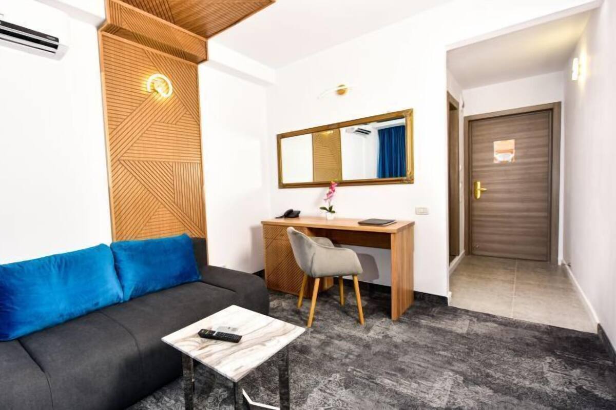 Et kompakt hotelværelse med en grå sofa, et lille træskrivebord, en stol, et spejl og en dør.