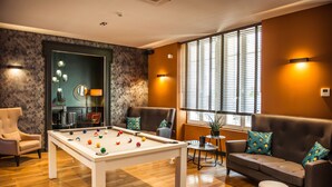 A pool table with balls, a sofa, and a chair in a room with wooden floors and a window with blinds.