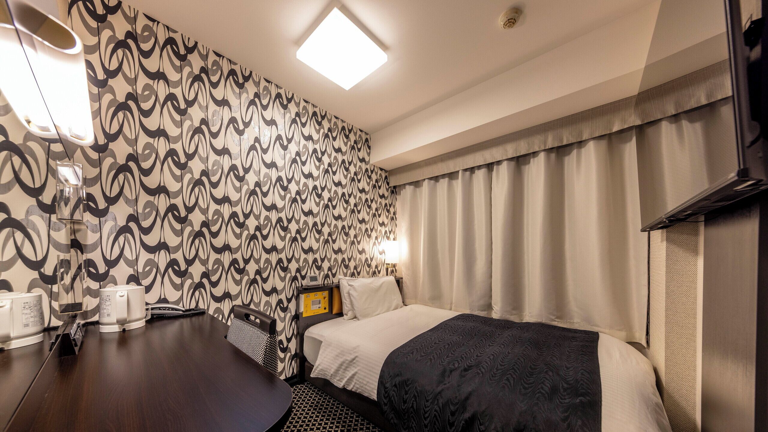 Una habitació d'hotel amb paper tapet motllejat, un llit amb llençol fosc, una tauleta de nit amb una làmpada i un televisor fixat a la paret.