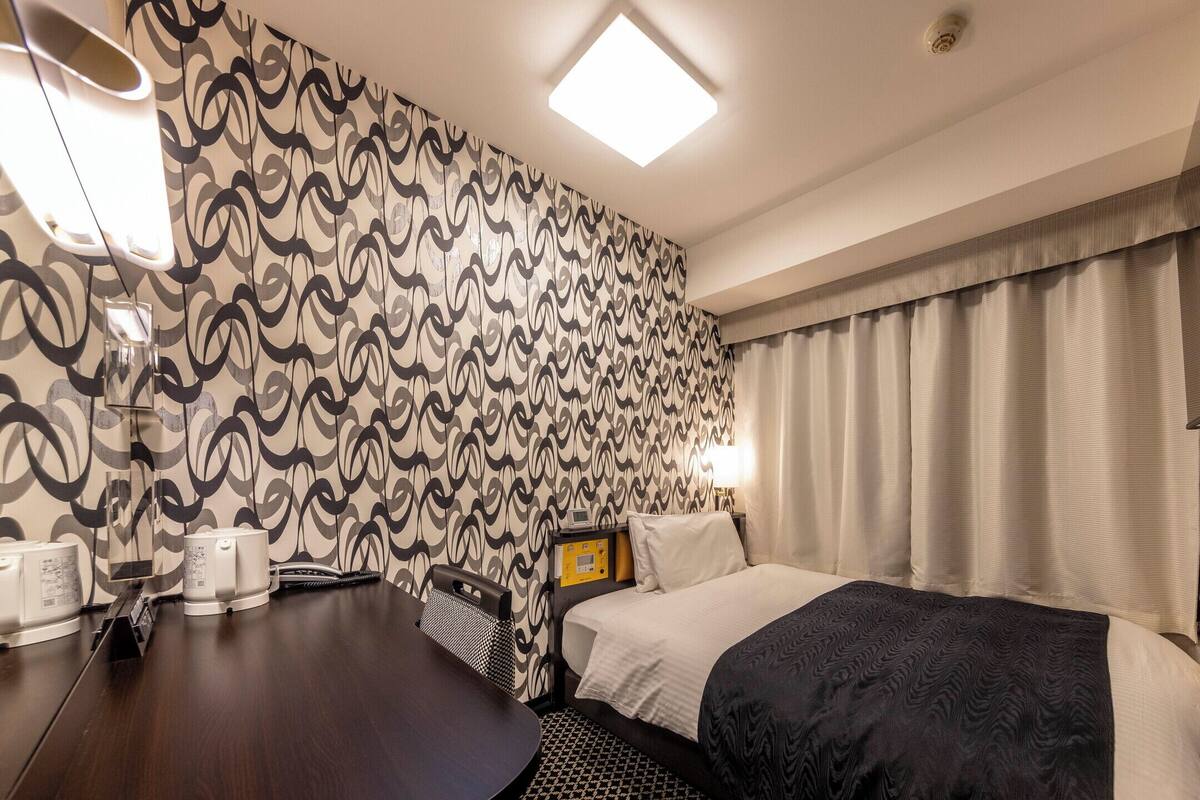 A hotel room with a patterned wallpaper, a bed with a dark blanket, a bedside table with a lamp, and a TV mounted on the wall.