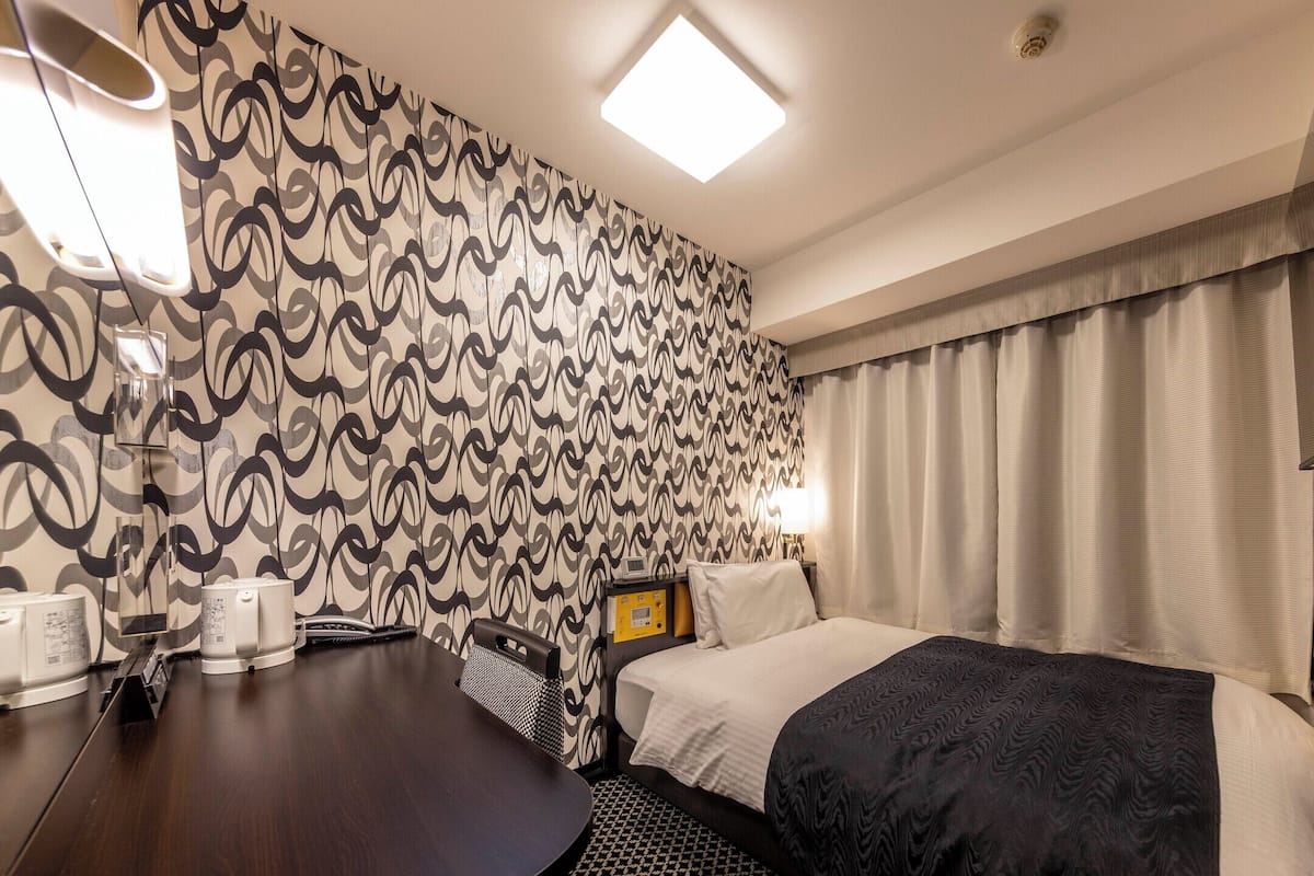 A hotel room with a patterned wallpaper, a bed with a dark blanket, a bedside table with a lamp, and a TV mounted on the wall.