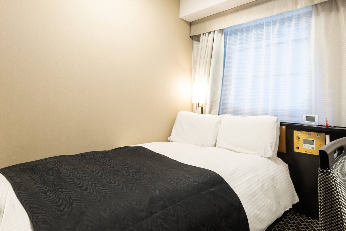 A hotel room with a bed, a bedside table with a lamp, and a window with curtains.