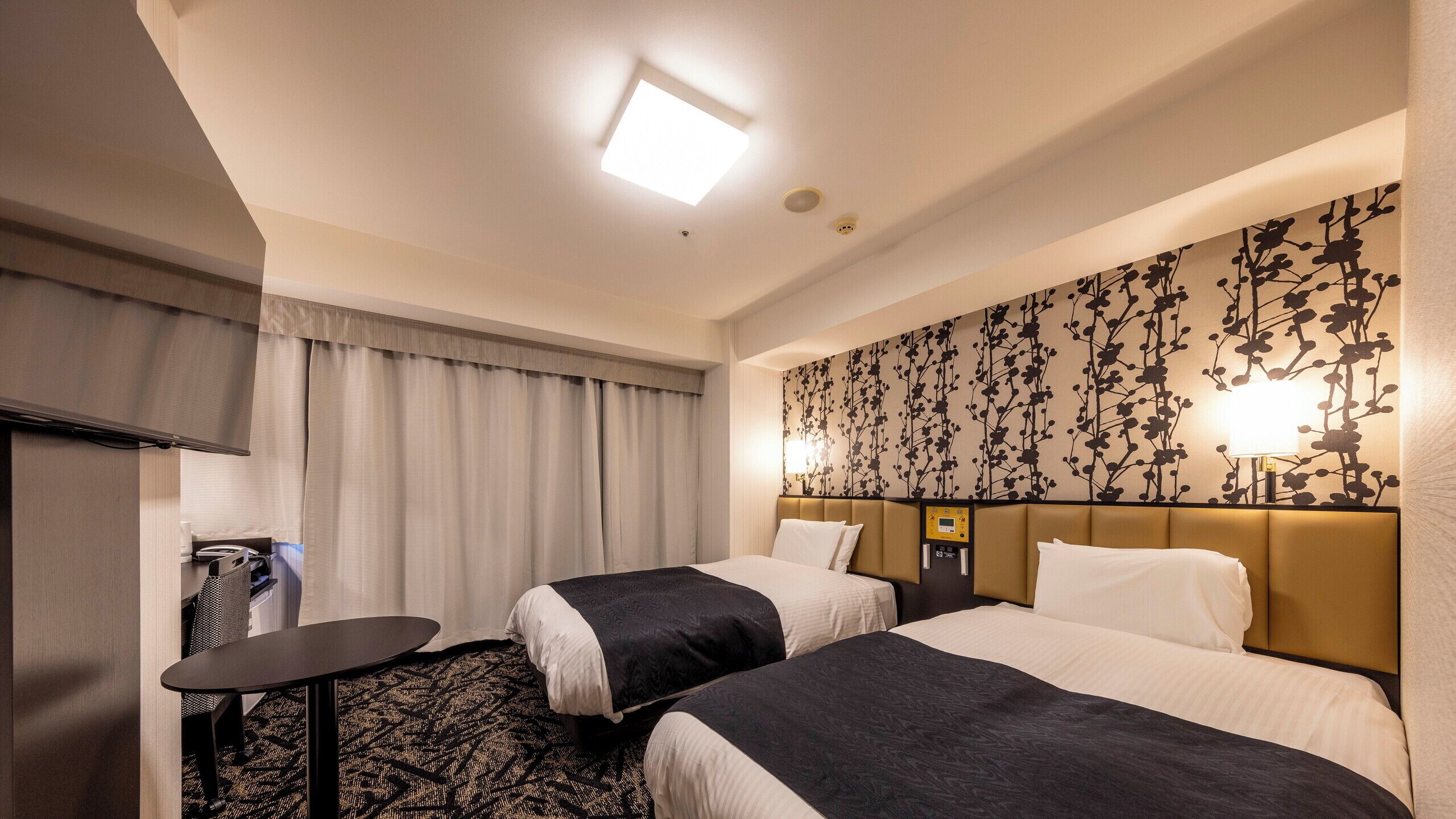 Habitació d'hotel amb dos llits, un escriptori, una cadira, una taula petita i una làmpada penjada a la paret.