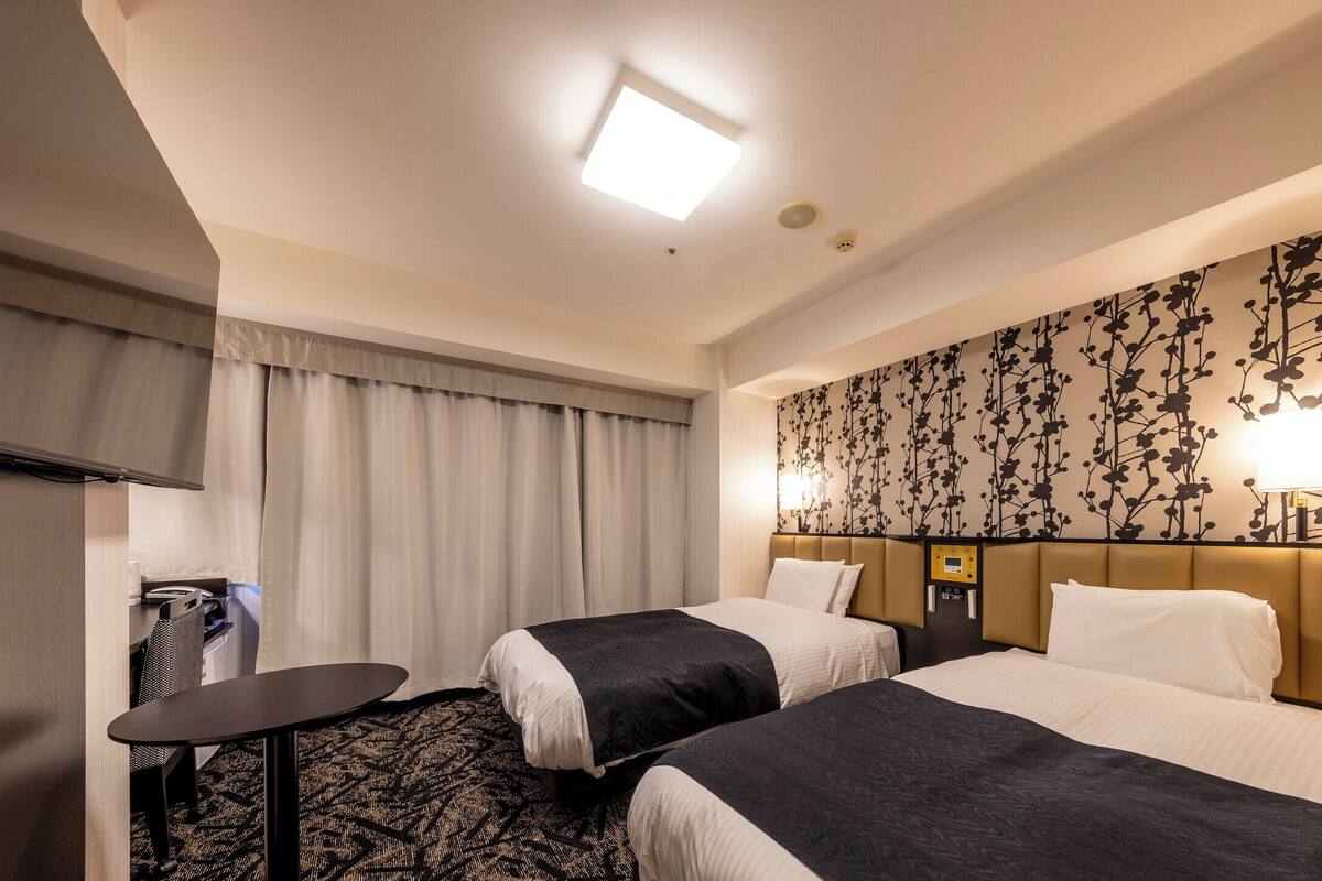 A hotel room with two beds, a desk, a chair, a small table, and a wall-mounted lamp.