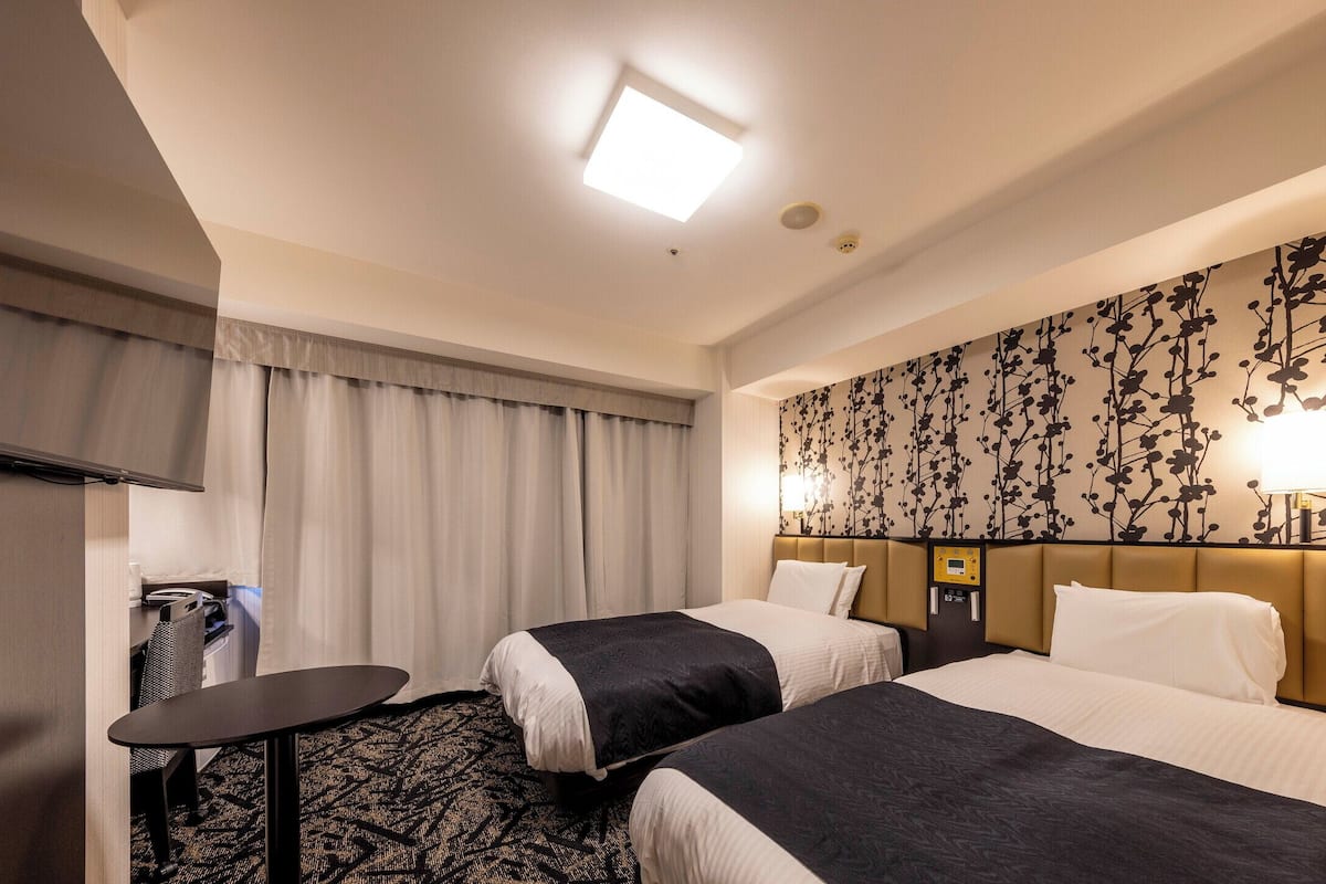 A hotel room with two beds, a desk, a chair, a small table, and a wall-mounted lamp.