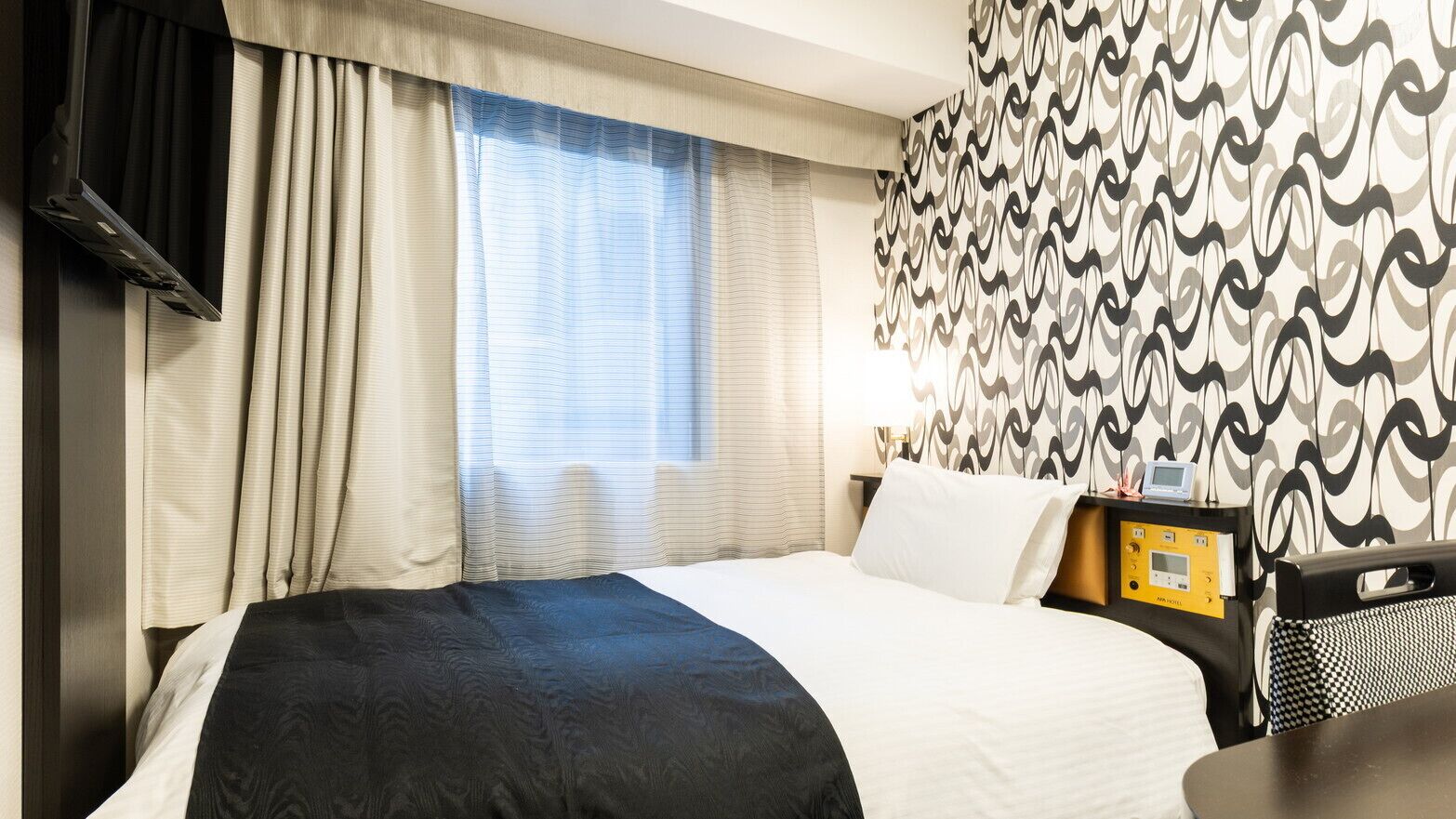 Ein Hotelzimmer mit Bett, Vorhängen, einer Wand mit Muster, einem Nachttisch mit Lampe und einem Fernseher.