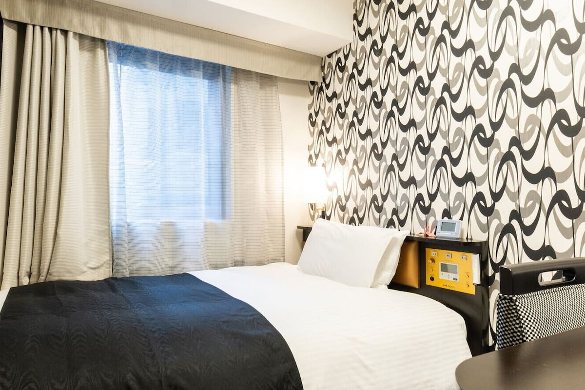 Ein Hotelzimmer mit Bett, Vorhängen, einer Wand mit Muster, einem Nachttisch mit Lampe und einem Fernseher.