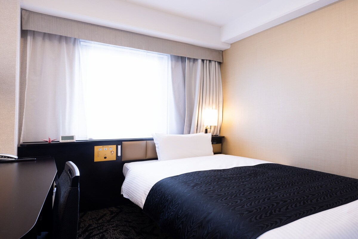 Habitación de hotel con cama, escritorio, silla, mesita de noche, lámpara y ventana con cortinas.