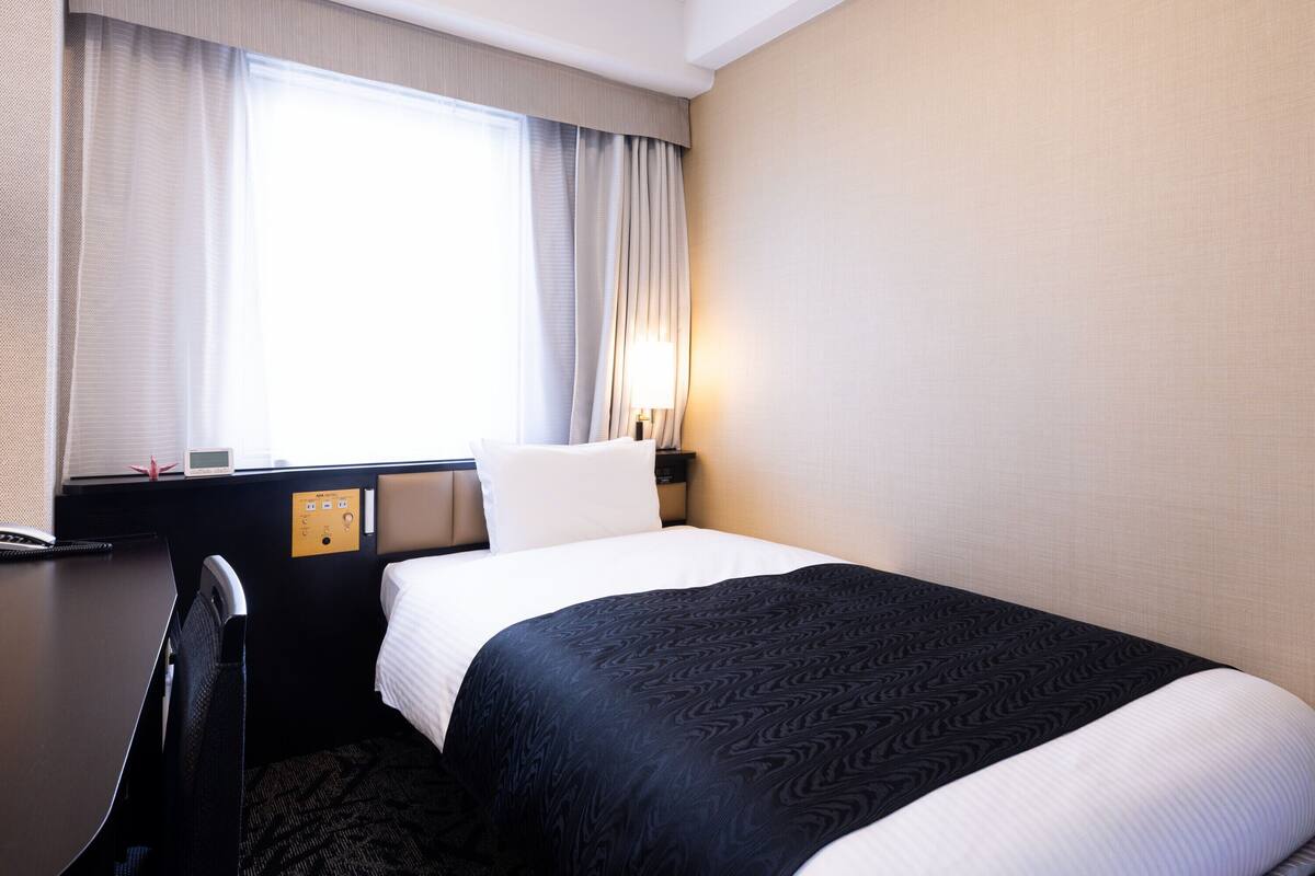 A hotel room with a bed, a desk, a chair, a window with curtains, and a wall-mounted lamp.