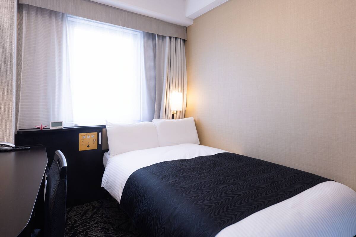 A hotel room with a bed, a desk with a lamp, a window with curtains, and a wall with a light switch.