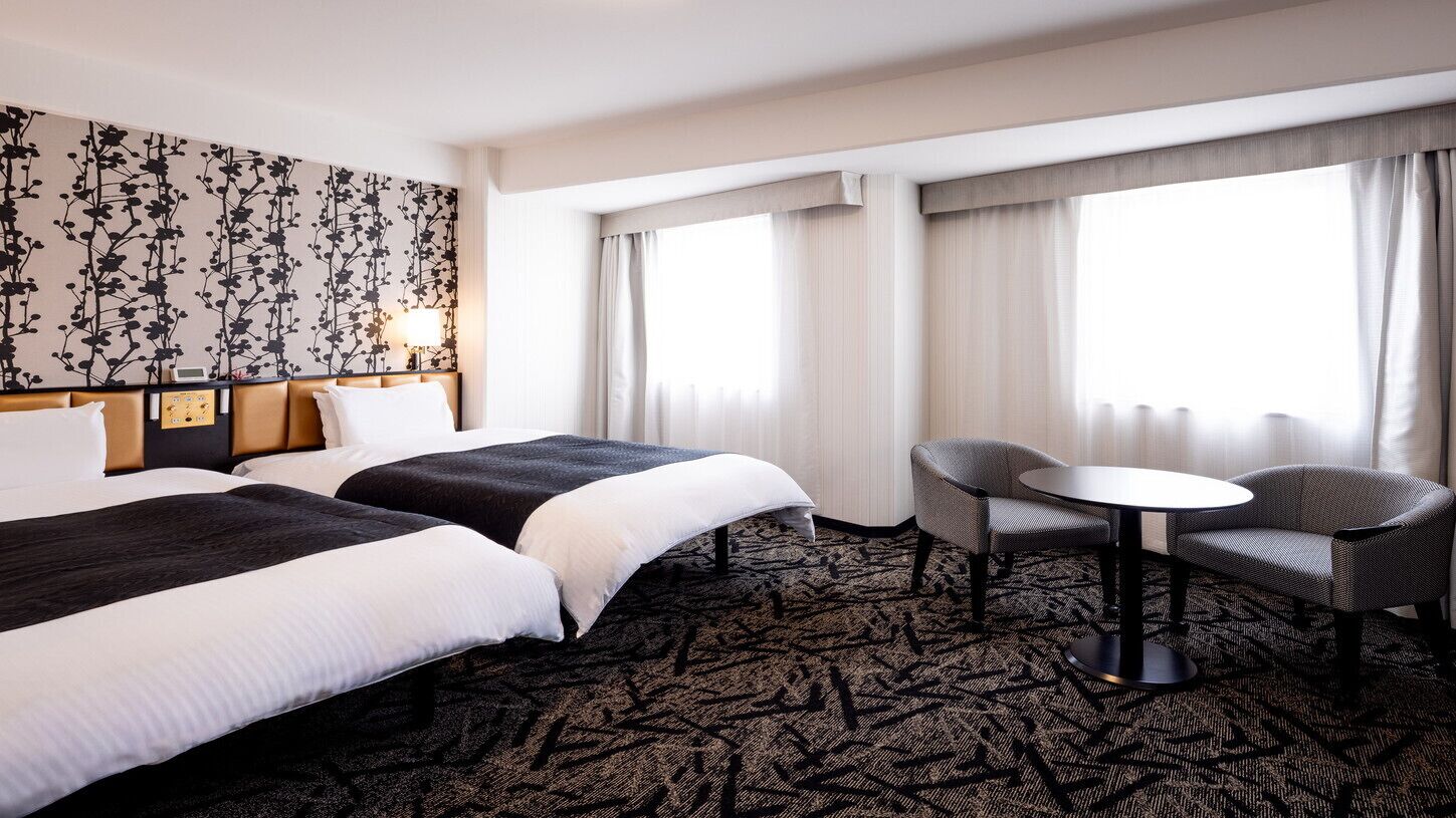 A hotel room with two beds, a small table, two chairs, a patterned carpet, and a large window with curtains.