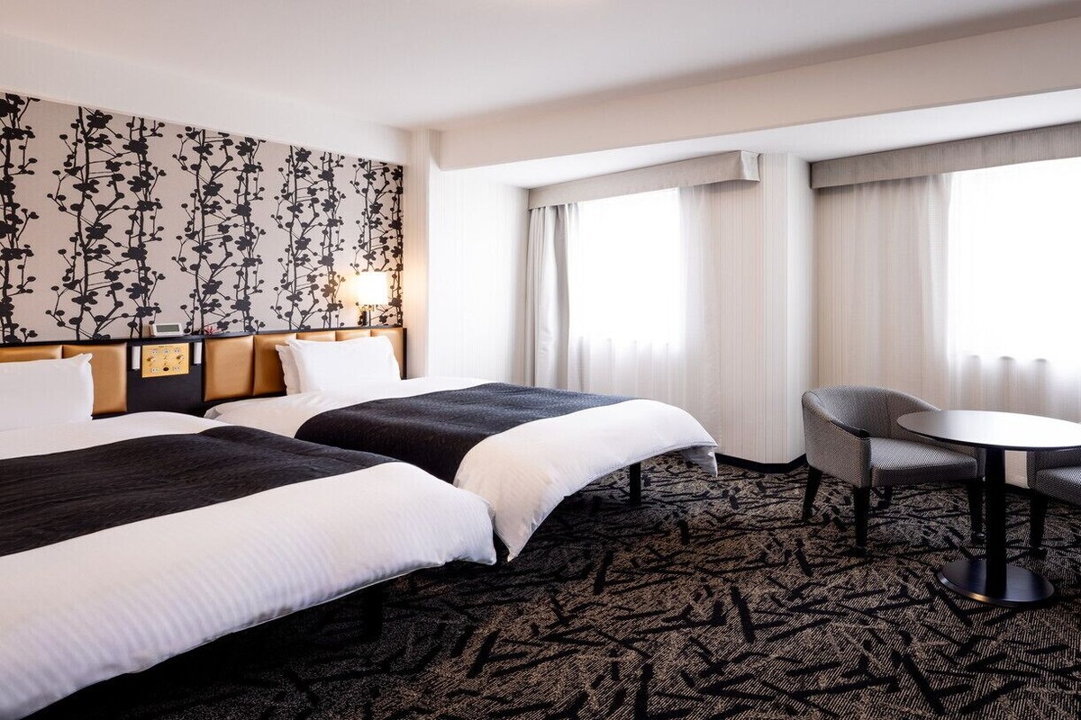 Habitación de hotel con dos camas, una mesita pequeña, dos sillas, alfombra estampada y un ventanal con cortinas.