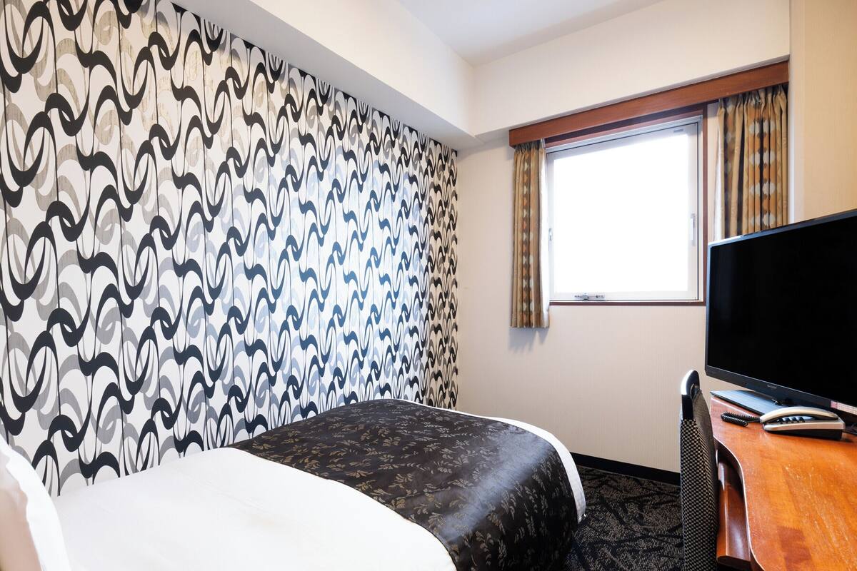 A hotel room with a patterned wall, a bed, a desk with a TV, and a window with curtains.