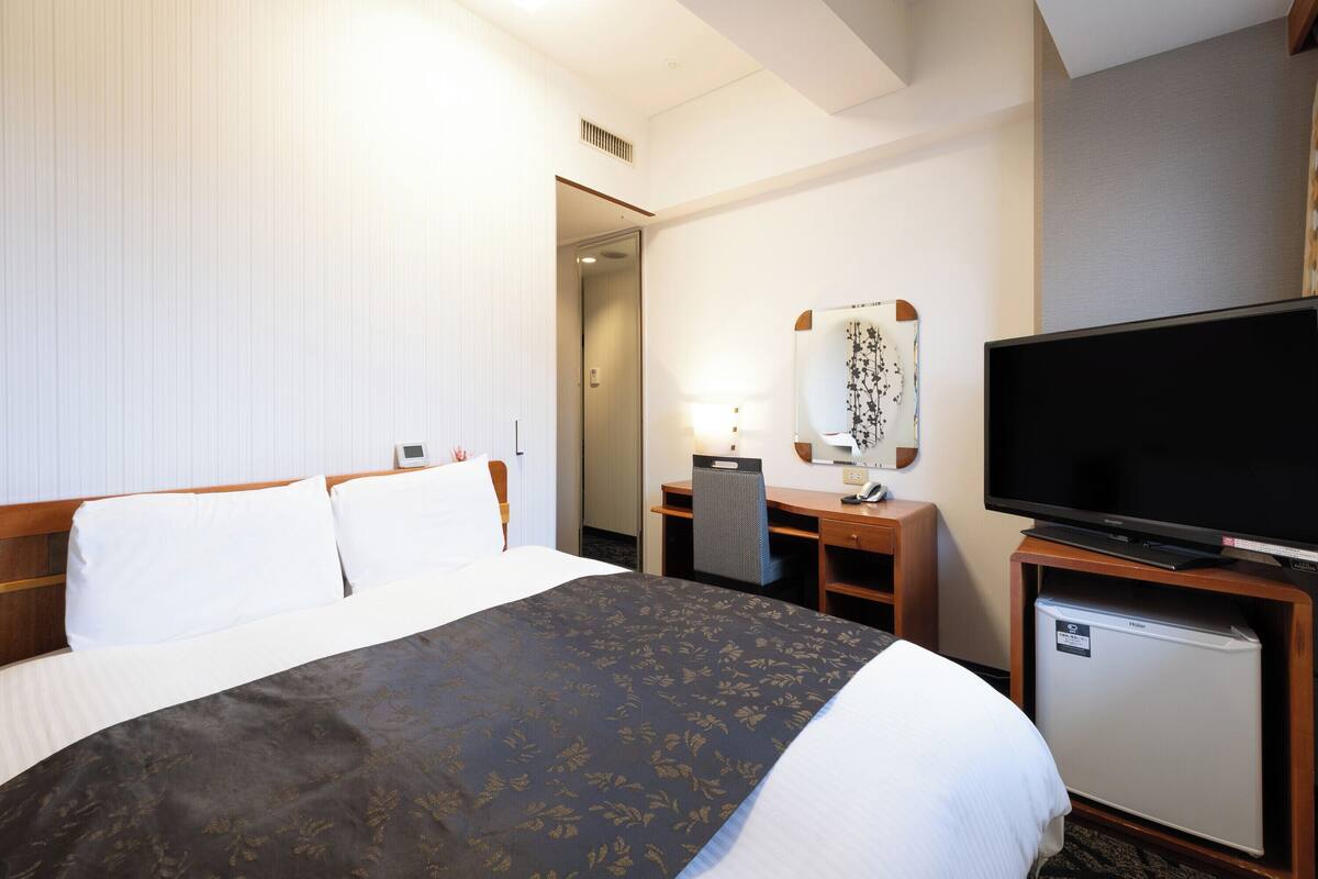 A hotel room with a bed, a desk with a chair, a television, and a mini-fridge.