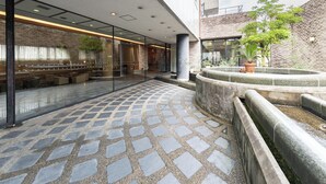 Un patio con suelo de piedra con diseño, un área de descanso con bancos y una pequeña fuente circular rodeada de plantas.