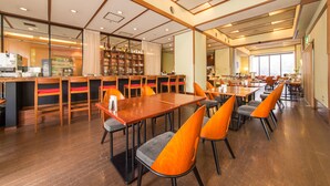 Un restaurante espacioso con mesas de madera y sillas color naranja, una zona de bar con estantes llenos de botellas y un ventanal que proporciona luz natural.