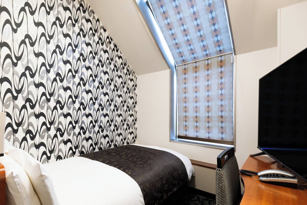 A hotel room with a bed, a desk with a telephone, and a window with patterned curtains.