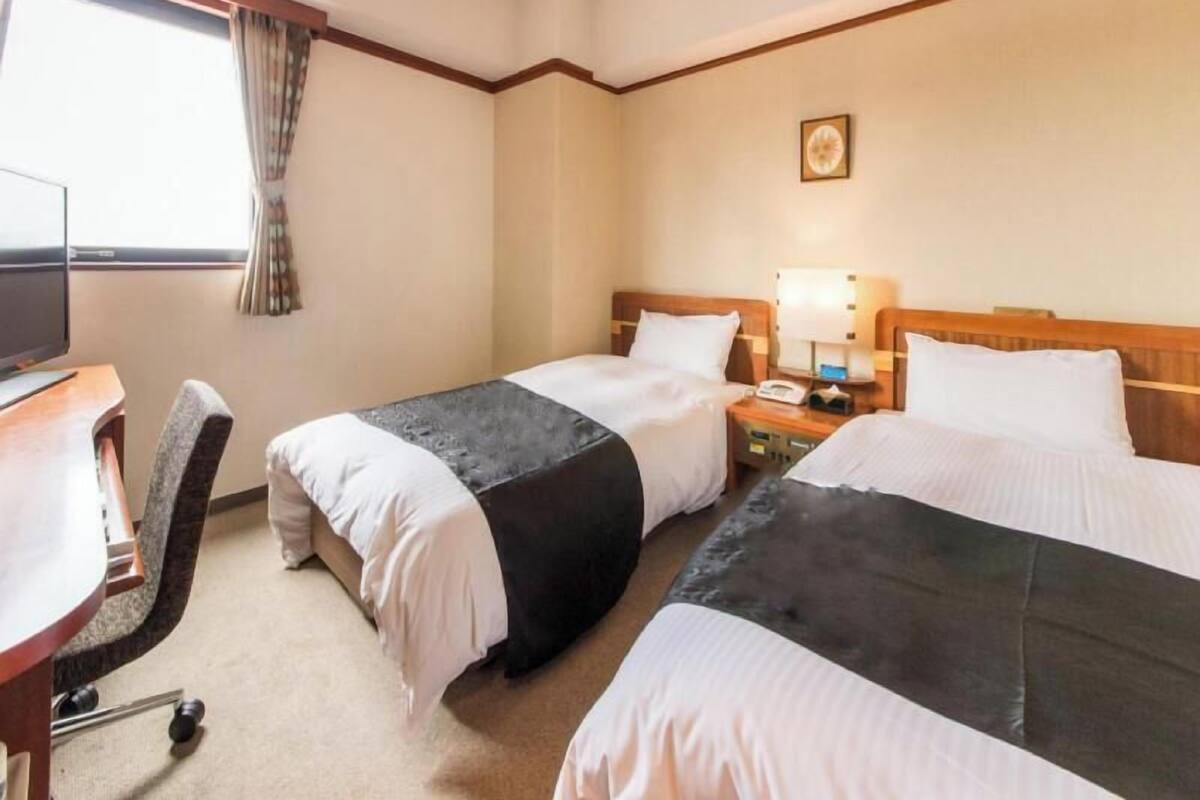 A hotel room with two beds, a desk with a computer, a chair, and a window with curtains.