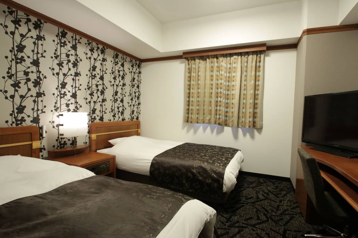 A hotel room with two beds, a TV, a nightstand, a lamp, and a window with curtains.