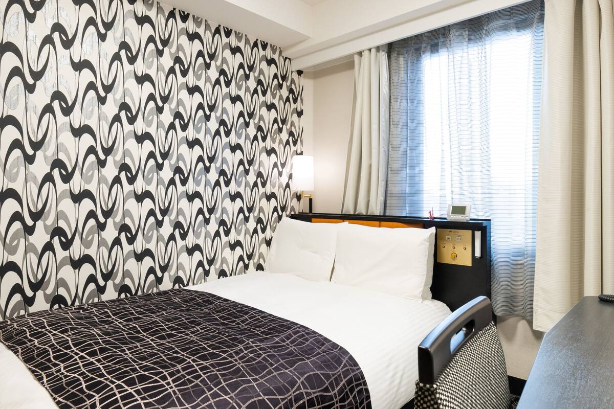 Ein Hotelzimmer mit strukturiertem Wandpapier, einem Bett mit weißen Leinen, einem Nachttisch mit Lampe, einer Steuerung und einem Fenster mit Vorhängen.