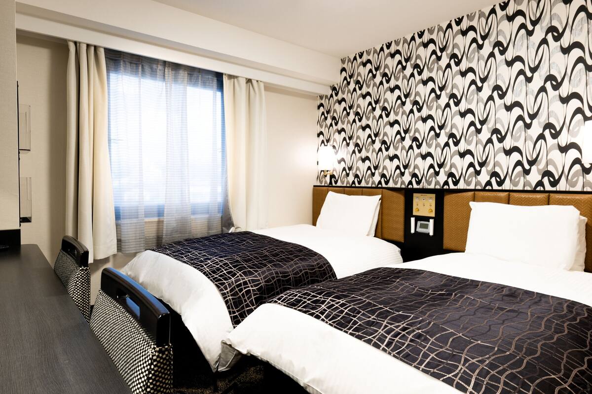 Ein Hotelzimmer mit zwei Betten, einer Wand mit Muster, einem Fenster mit Vorhängen und einer Bedienkonsole an der Wand.