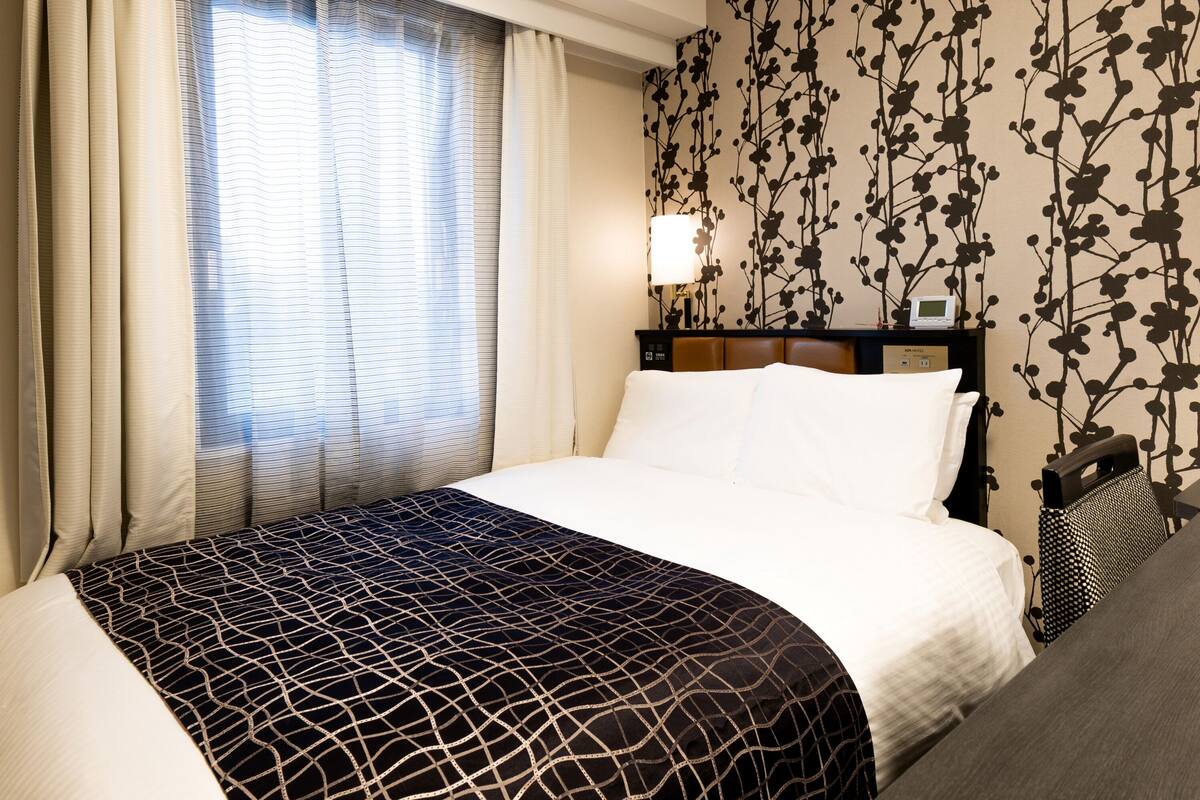 Ein Hotelzimmer mit einem Bett, Vorhängen, einer Wandlampe und einem blumigen Wandpapier-Muster.
