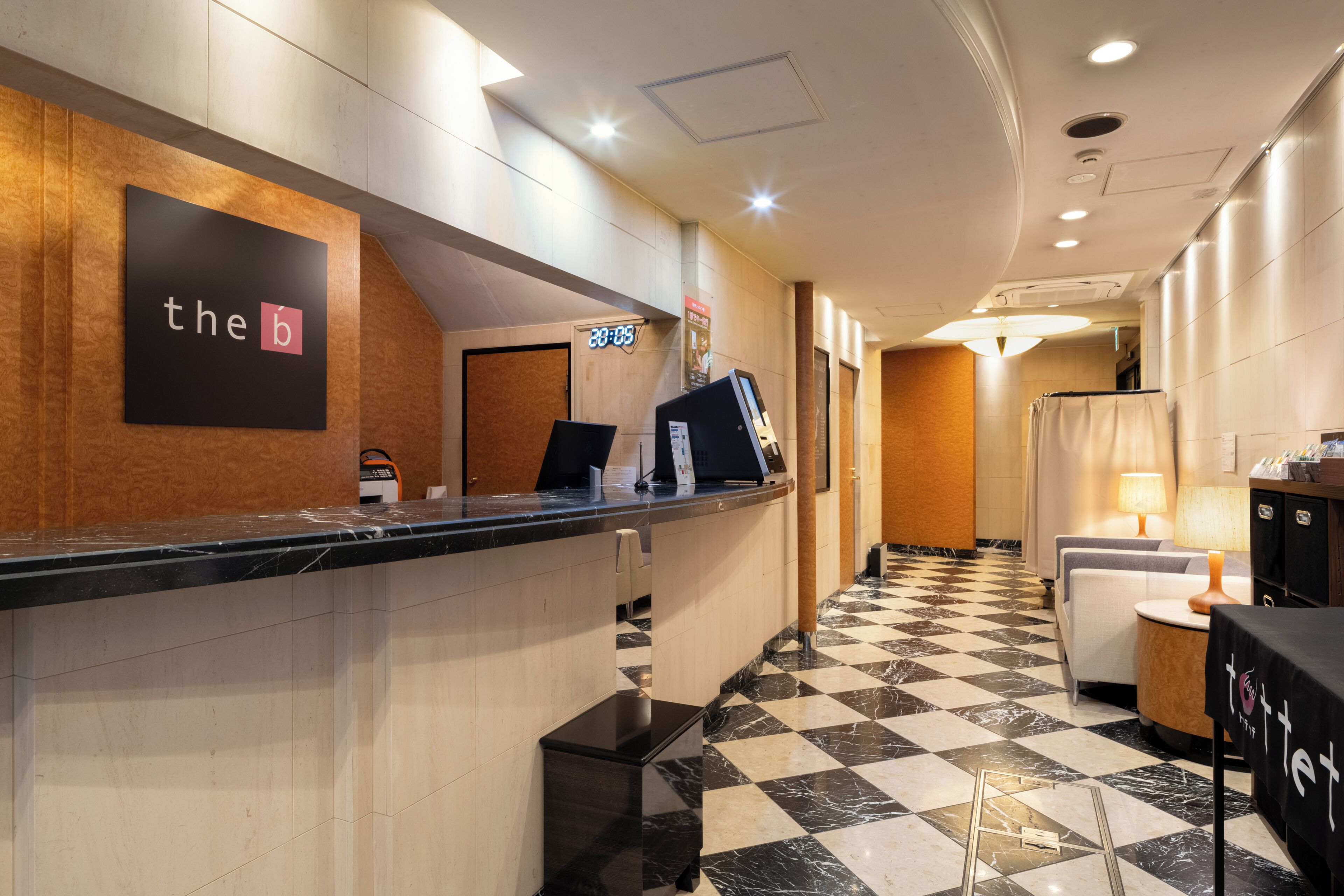 A hotel reception area with a check-in counter, a clock displaying 3:05, a reception desk, a seating area with a lamp, and a check-in counter with a sign reading 'the b'.